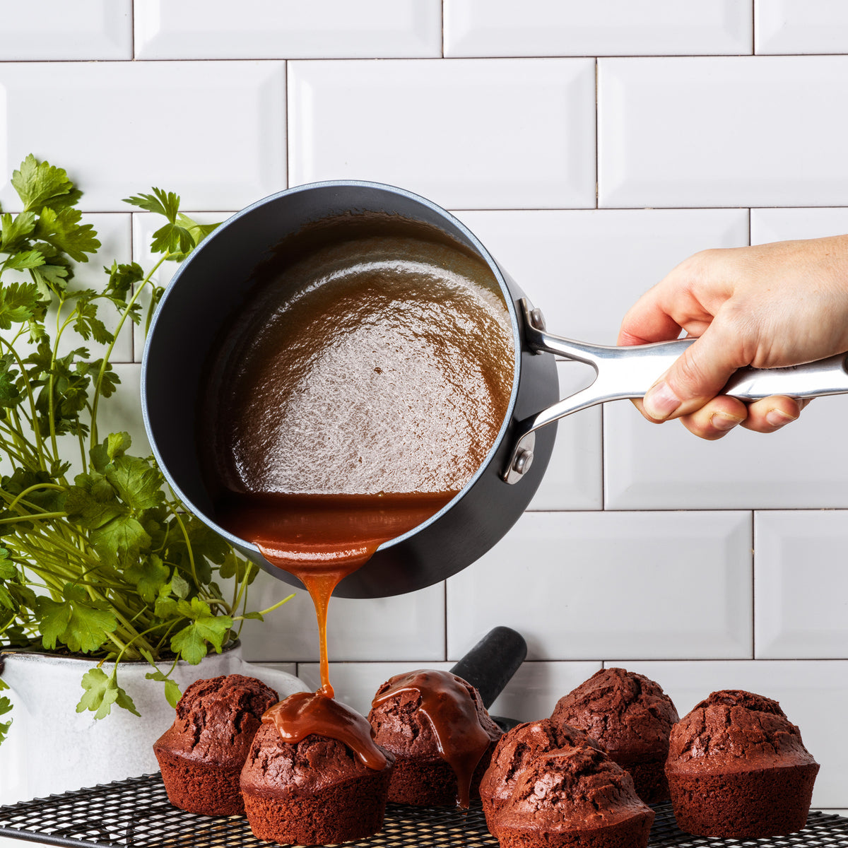 Caramel sauce is poured from the Valencia Pro Ceramic Nonstick 1.5-Quart Saucepan onto chocolate muffins on a wire rack, with fresh green herbs in a white pot and a white tile wall in the background.