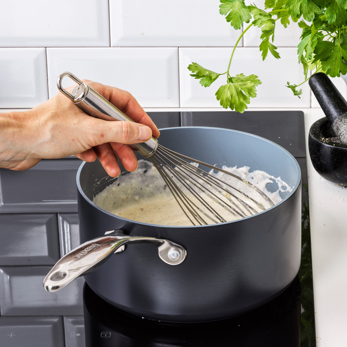 A hand stirs a creamy white sauce with a metal whisk in the Valencia Pro Ceramic Nonstick 1.5-Quart Saucepan on a stovetop, with fresh parsley and a mortar and pestle nearby.