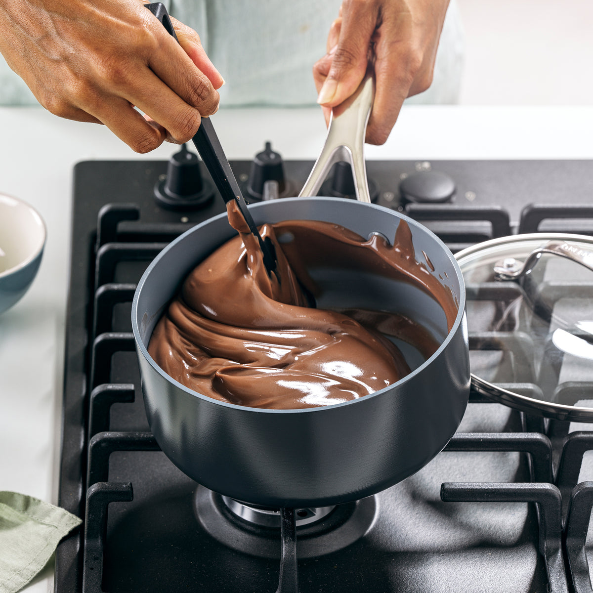 Hand stirring melted chocolate in a grey ceramic nonstick saucepan with lid on a stovetop.