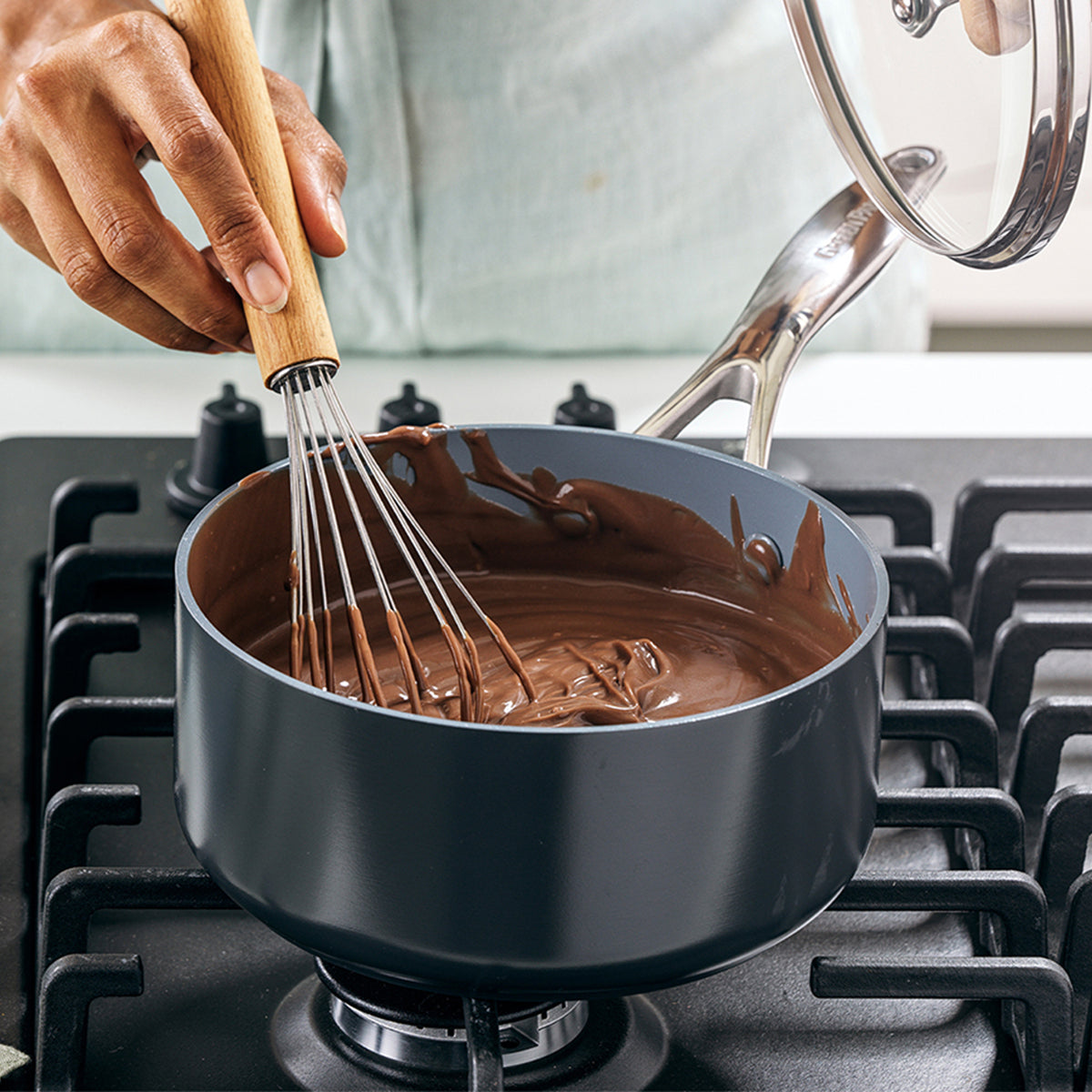 Melted chocolate being whisked in a gray ceramic nonstick 2-quart saucepan with lid on a stovetop.