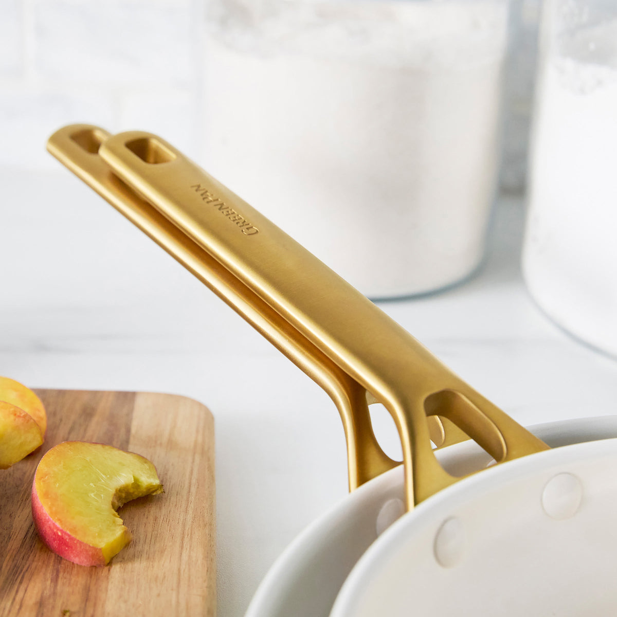 Cream frypans with gold handles on a white surface with apples and a cutting board.