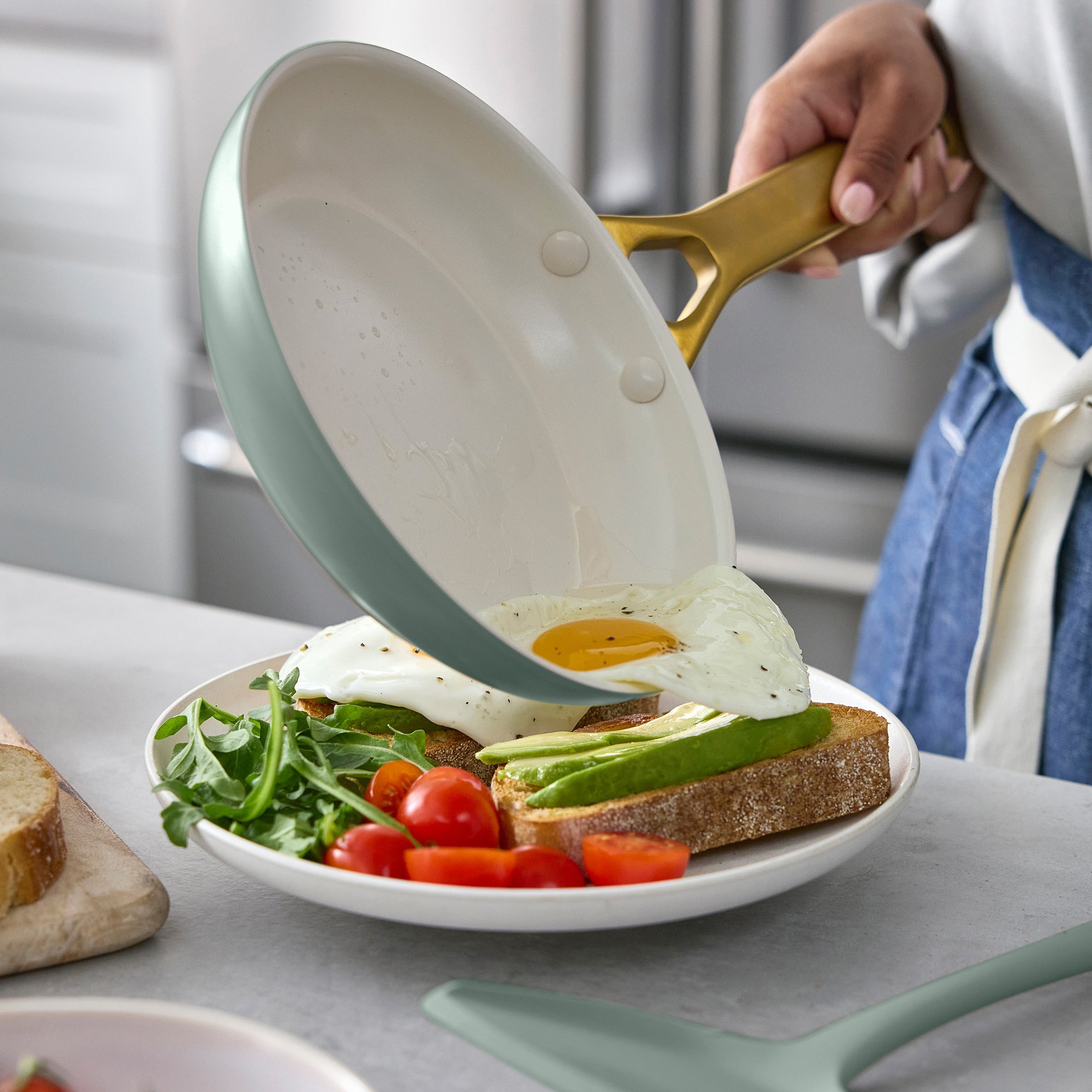 Person holding a green frying pan with an egg over a plate of toast, salad, and tomatoes.