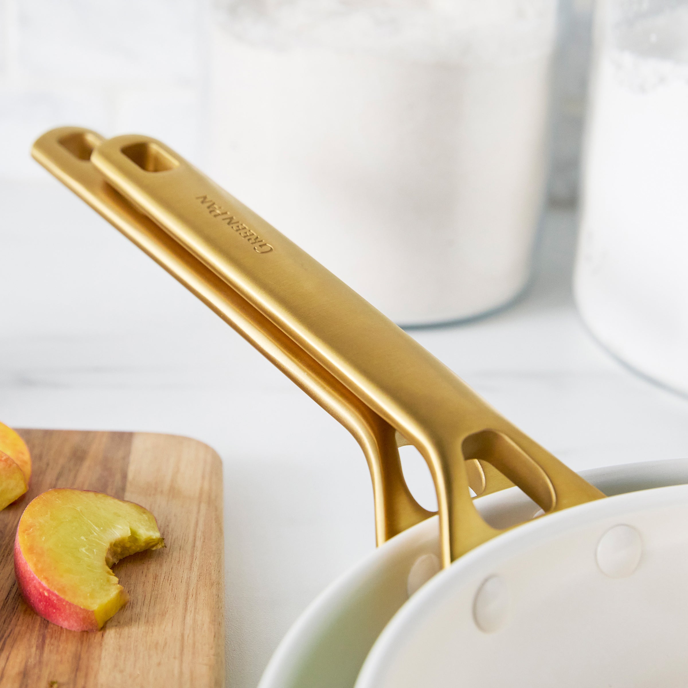 Green frypans with gold handles on a white surface with apples and a cutting board.