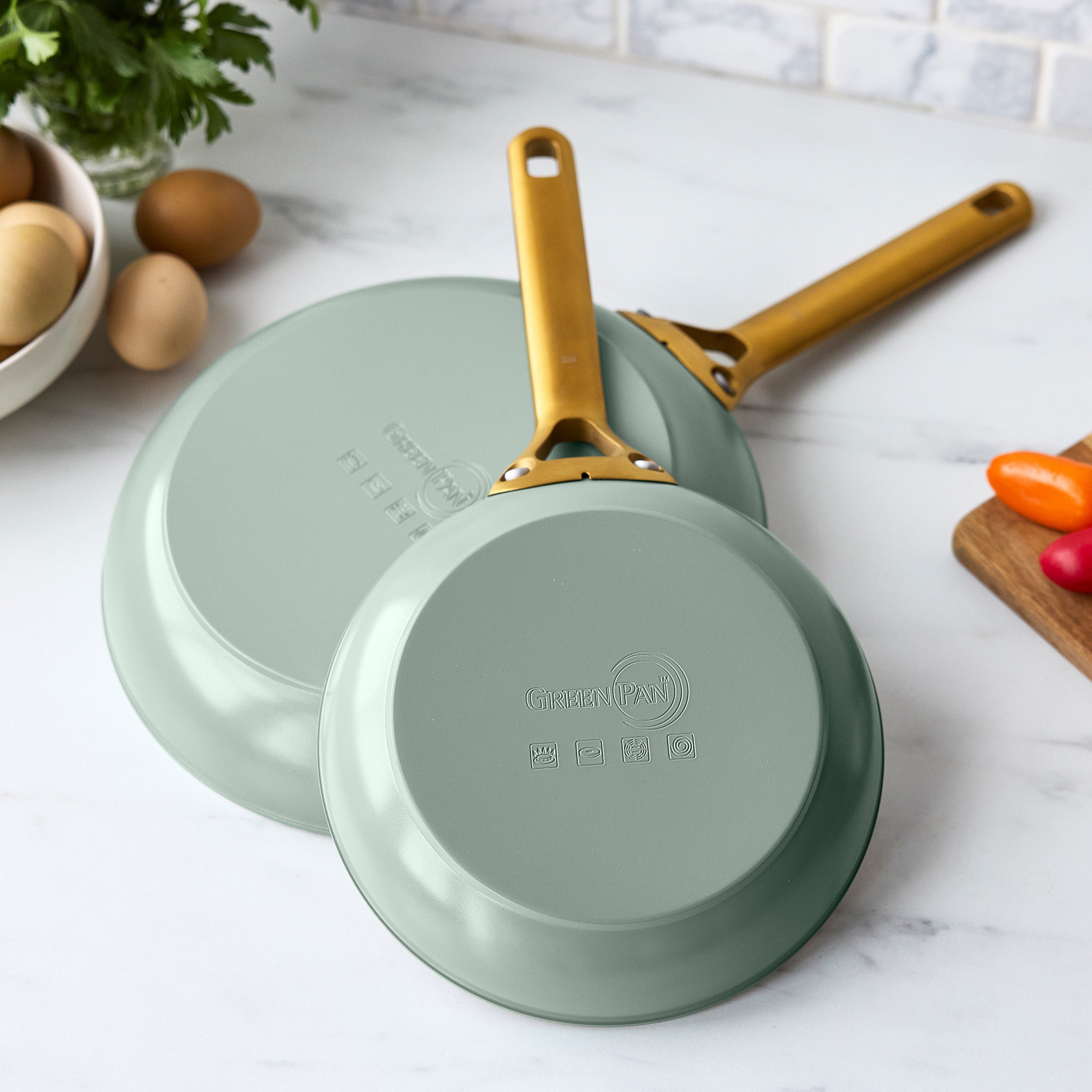 Two green frying pans with gold handles on a white surface.