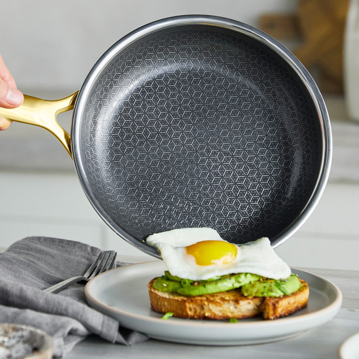 Gold ceramic nonstick frypan held over a plate with avocado toast and a sunny-side-up egg.
