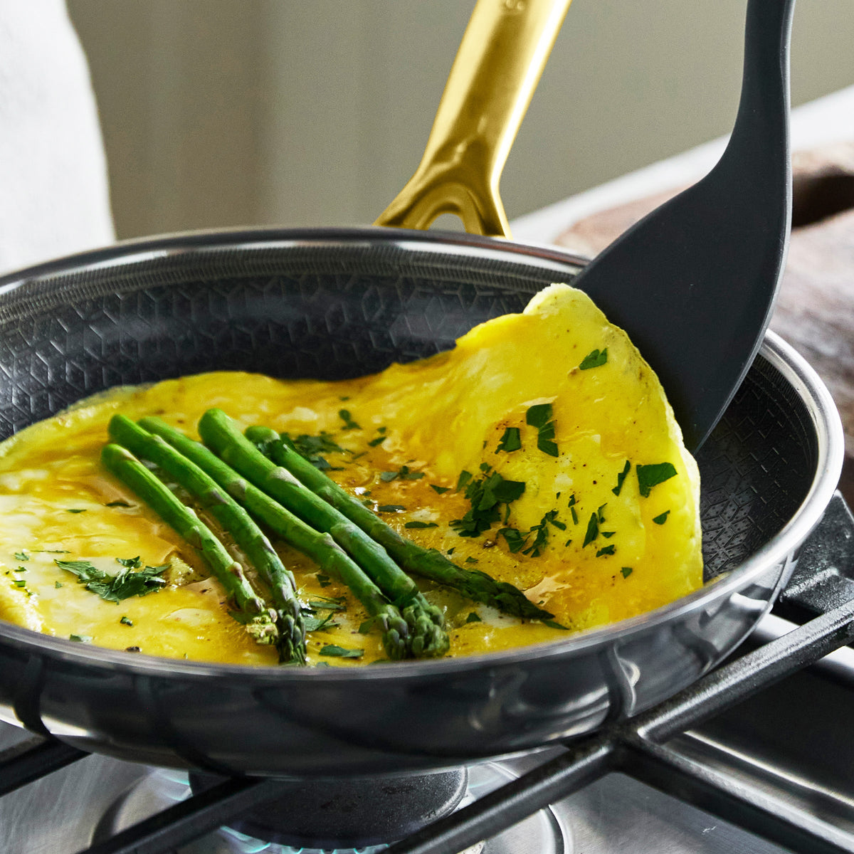 Omelette with asparagus and herbs being folded in a gold ceramic nonstick frypan on a stove.
