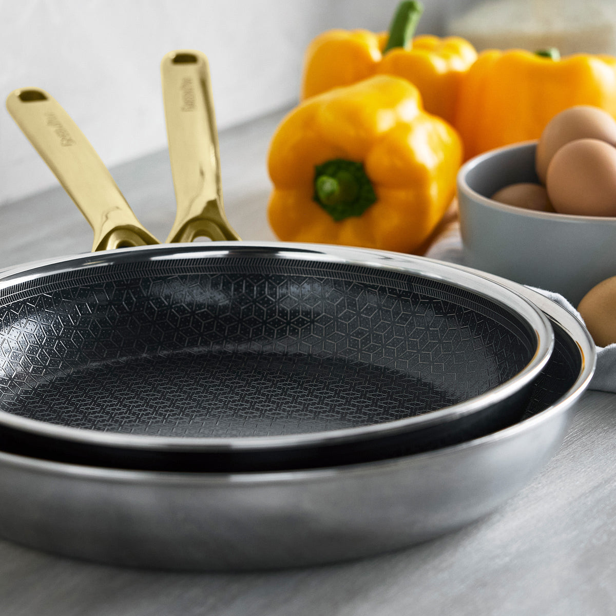 Gold ceramic nonstick frypan set of two, 10 and 12, shown beside bell peppers and brown eggs.