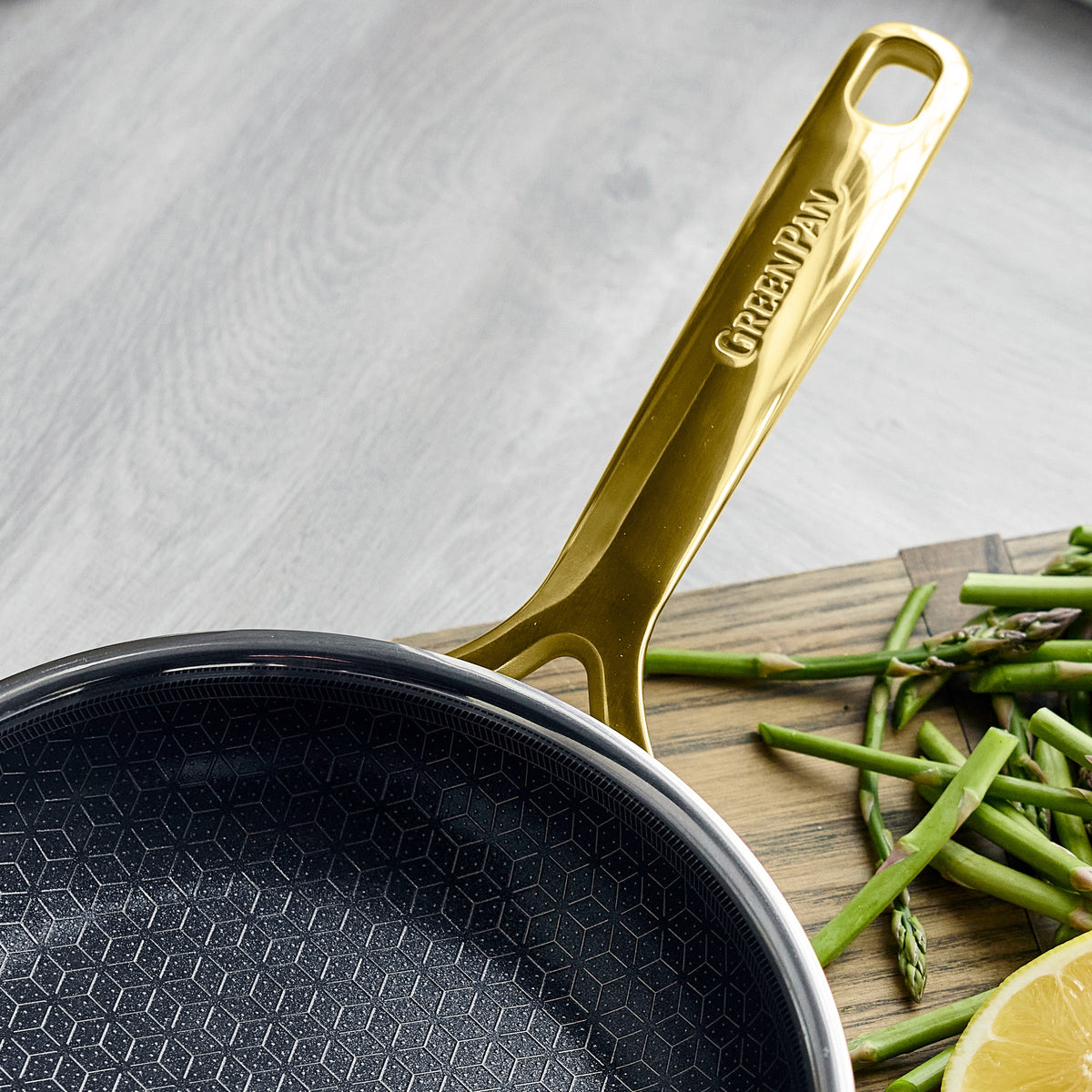 Gold ceramic nonstick frypan set on a countertop with chopped asparagus and a lemon wedge.