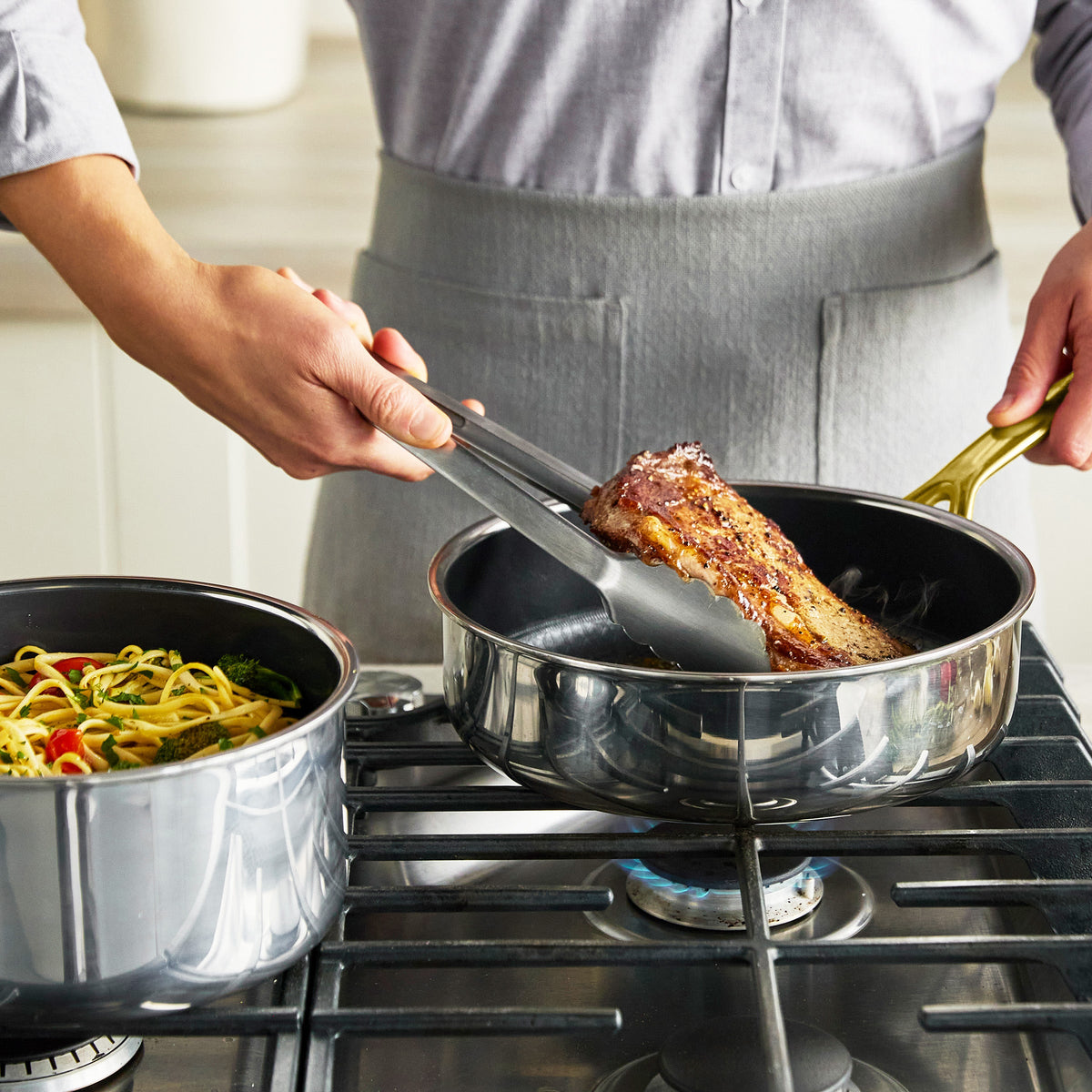 Steak cooking in a gold ceramic nonstick pan on the stove, with pasta simmering in a pot beside it.