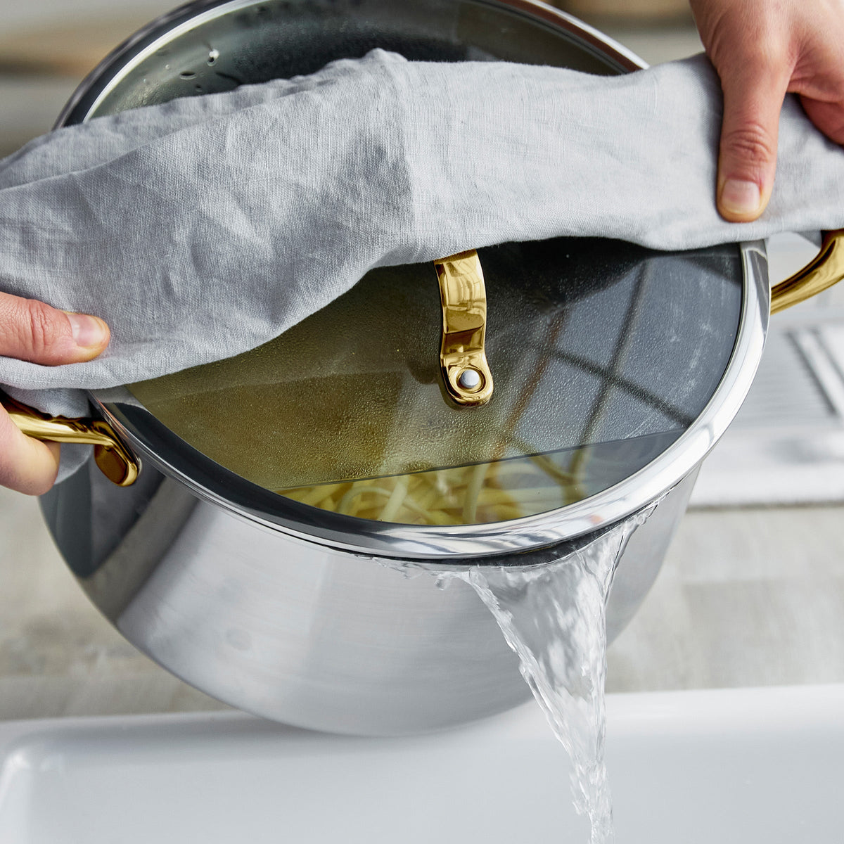 Hands holding a gold ceramic nonstick pot, pouring pasta water into a kitchen sink.