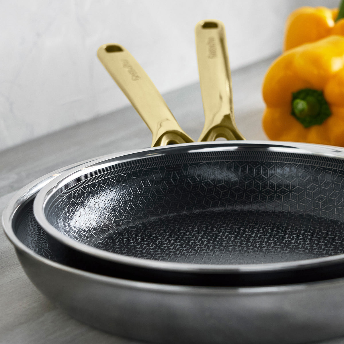Two gold hybrid pans on a counter with two yellow bell peppers nearby.