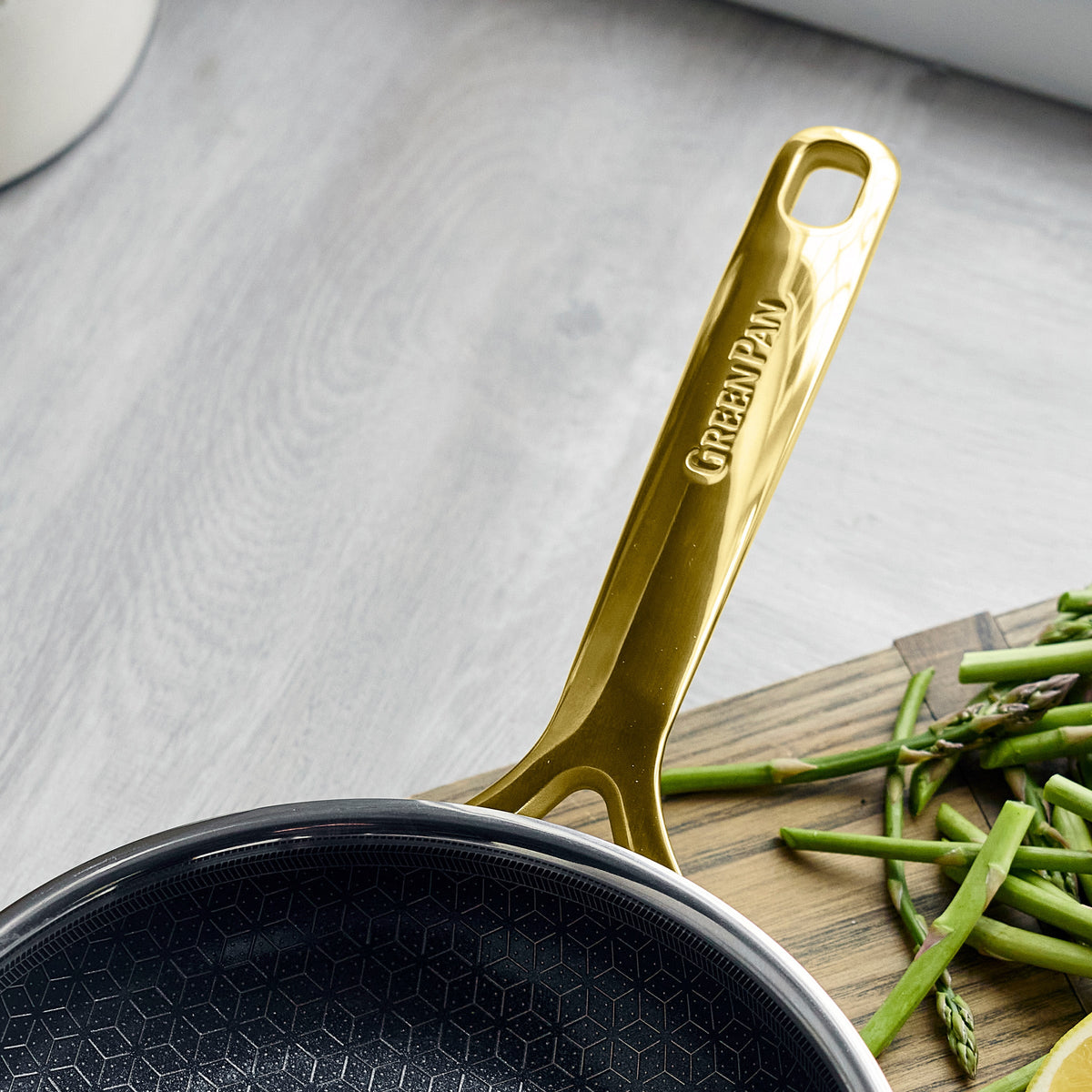 Gold ceramic nonstick frying pan from a 10-piece set next to fresh asparagus on a wooden board.