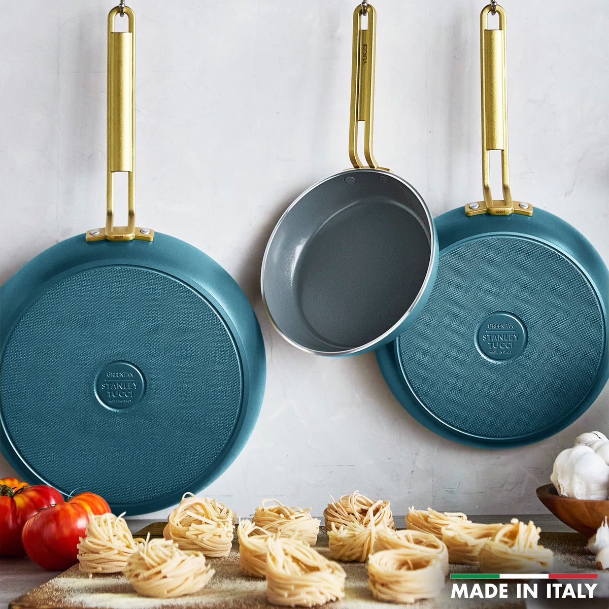 Three teal frying pans with gold handles on a white wall, with pasta and tomatoes on a wooden board below.