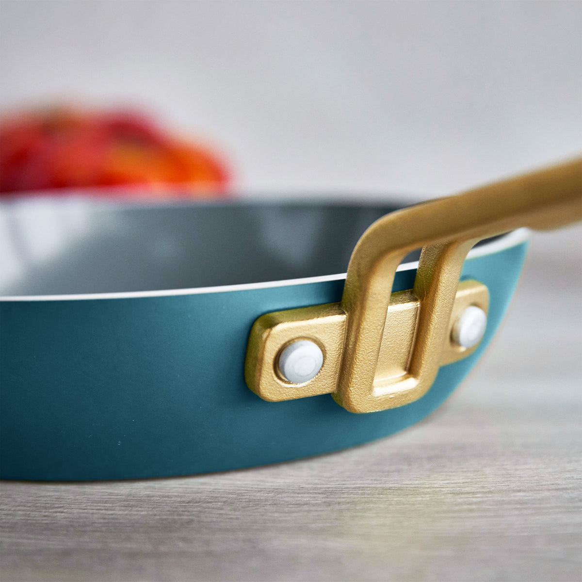 Close-up of a teal frying pan with a gold handle on a light wooden surface.