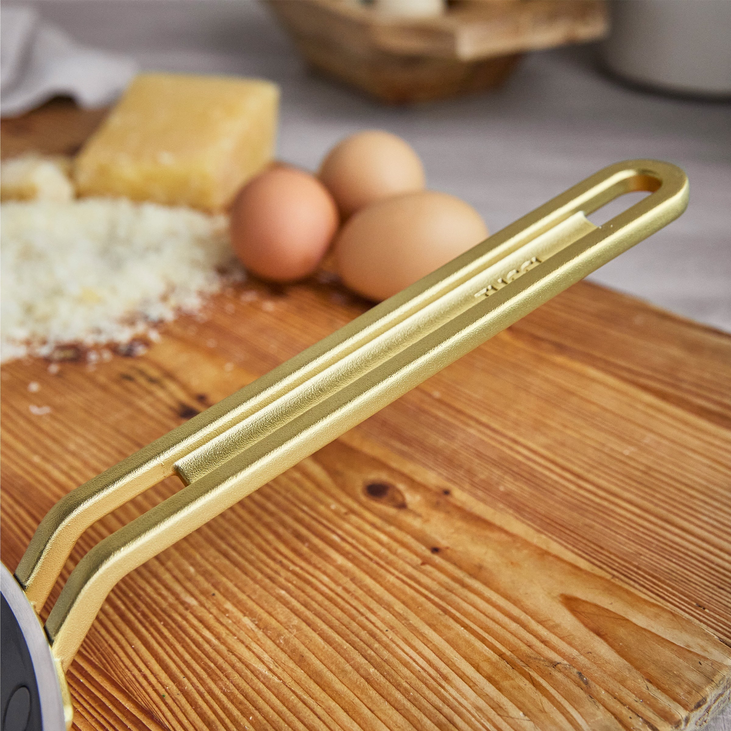 Gold crepe pan handle on a wooden cutting board with eggs and cheese in the background
