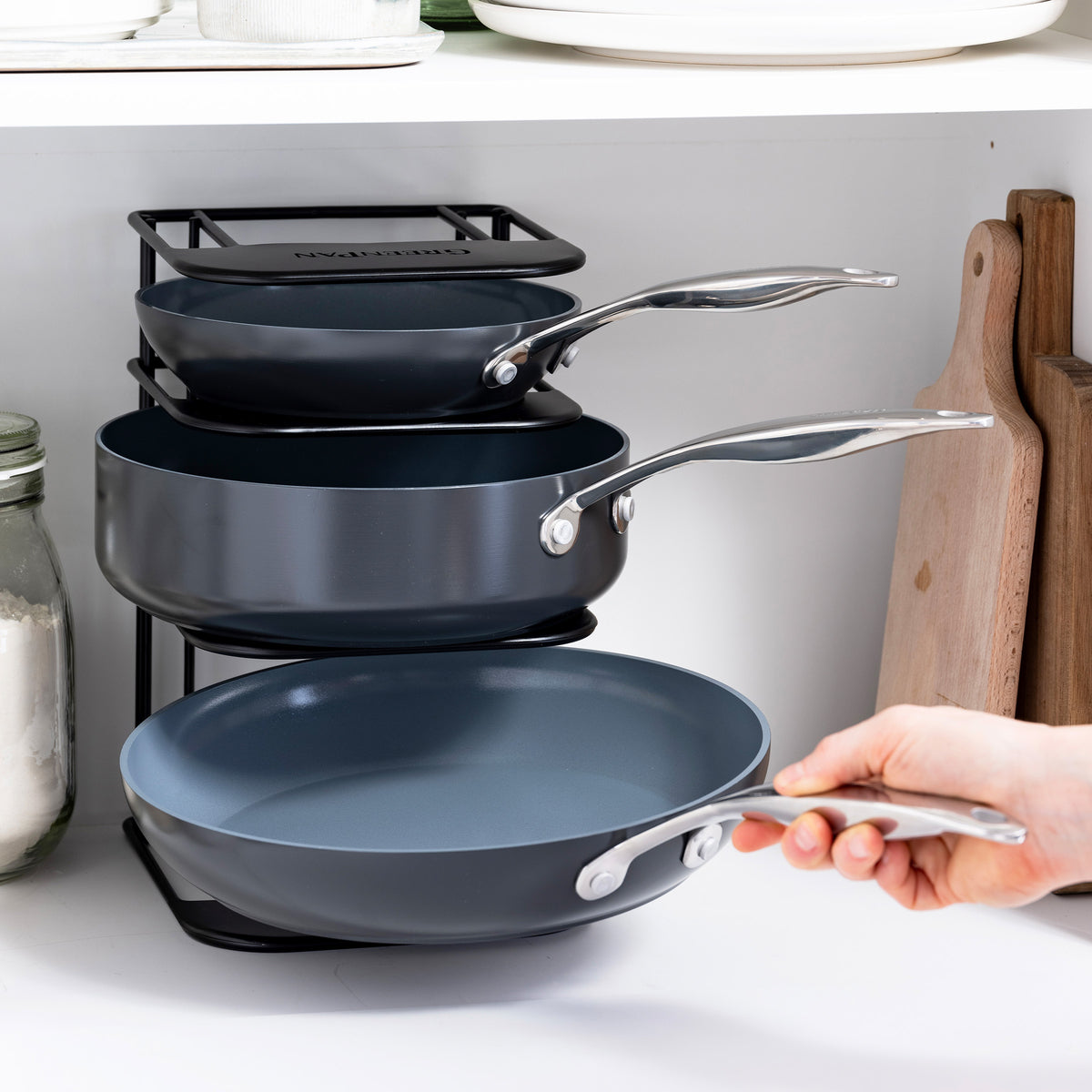 Hand pulling the bottom pan from a three-piece GreenPan cookware rack organized inside a cabinet.