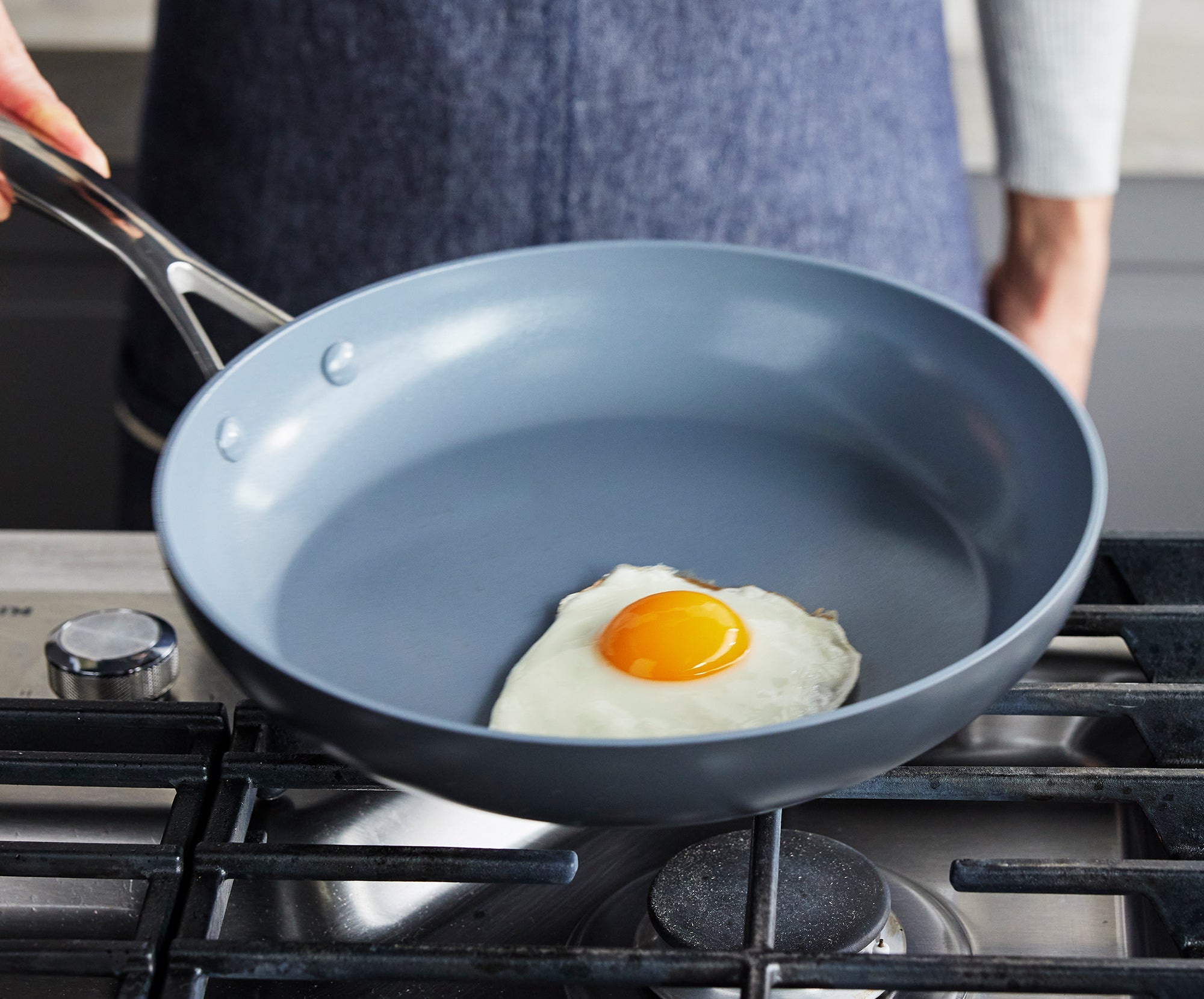 Hand holding a GreenPan frying pan with a single sunny side up egg cooking on a gas stove burner.