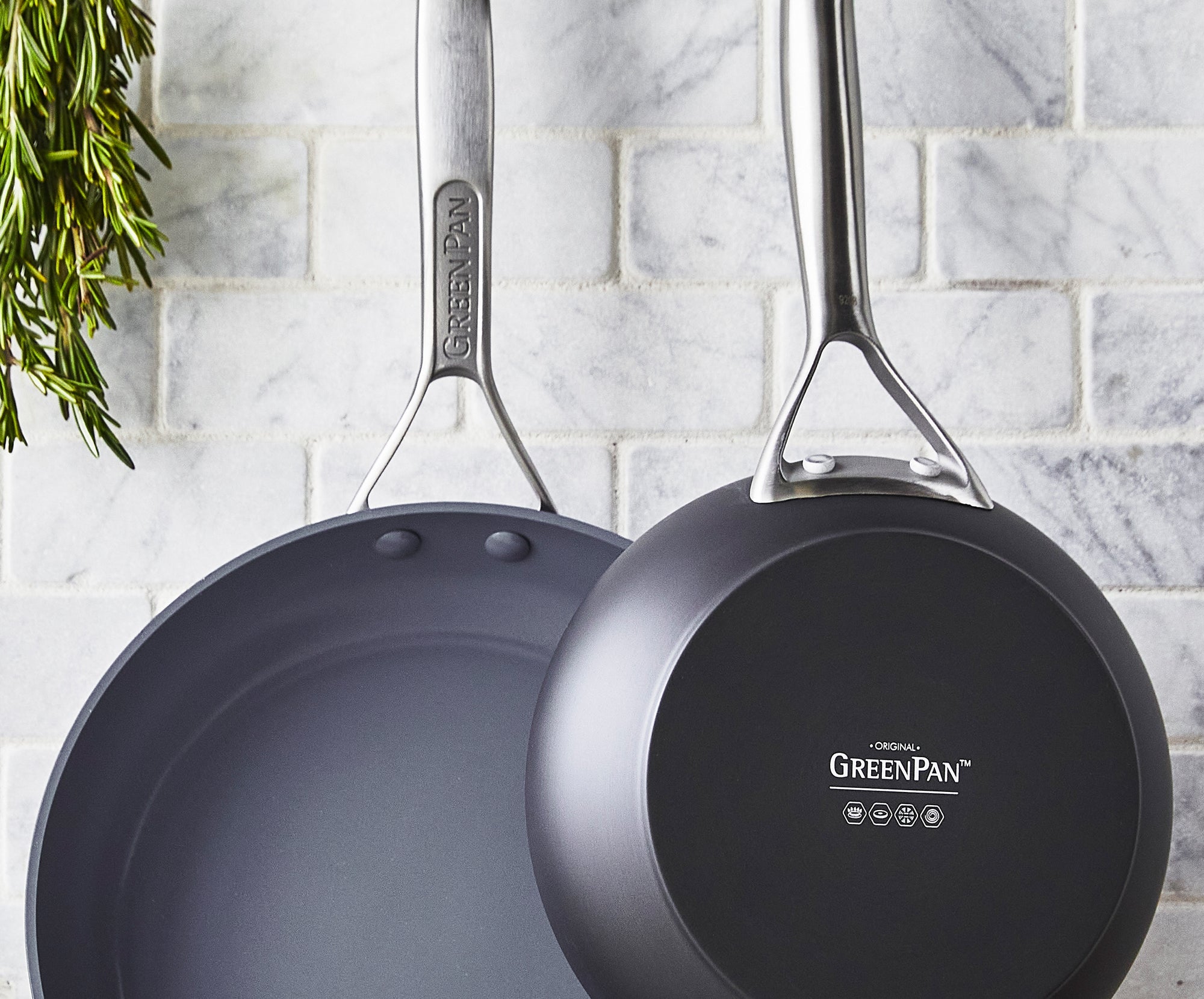 Two green nonstick skillets hang on a white tiled wall, with a sprig of fresh rosemary visible on the left.