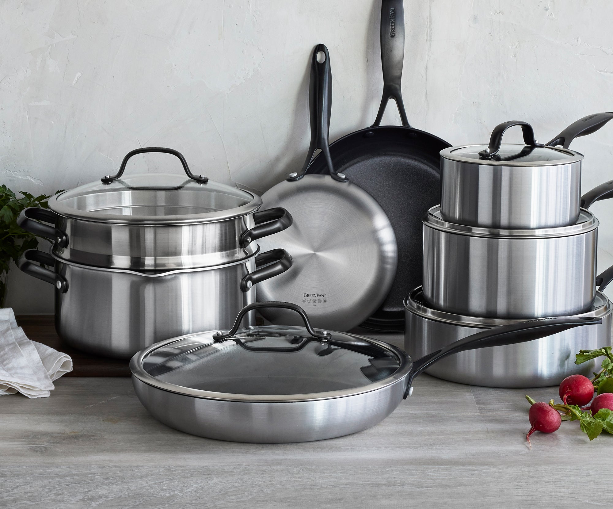 GreenPan stainless steel cookware set with lids on a kitchen counter, near radishes and a towel.