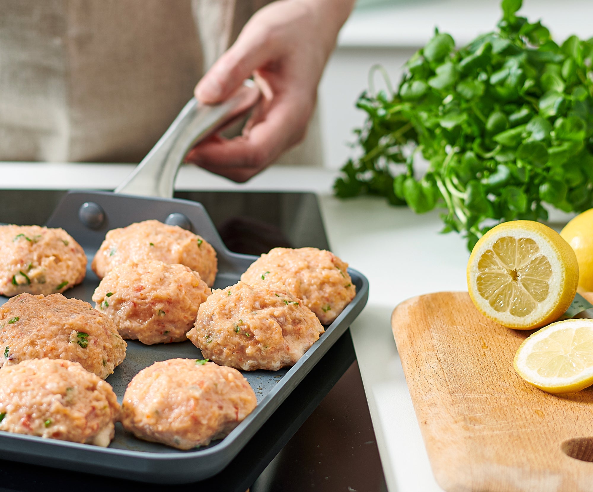 Raw chicken patties in a white ceramic nonstick pan; fresh herbs and lemon on a nearby wooden cutting board.