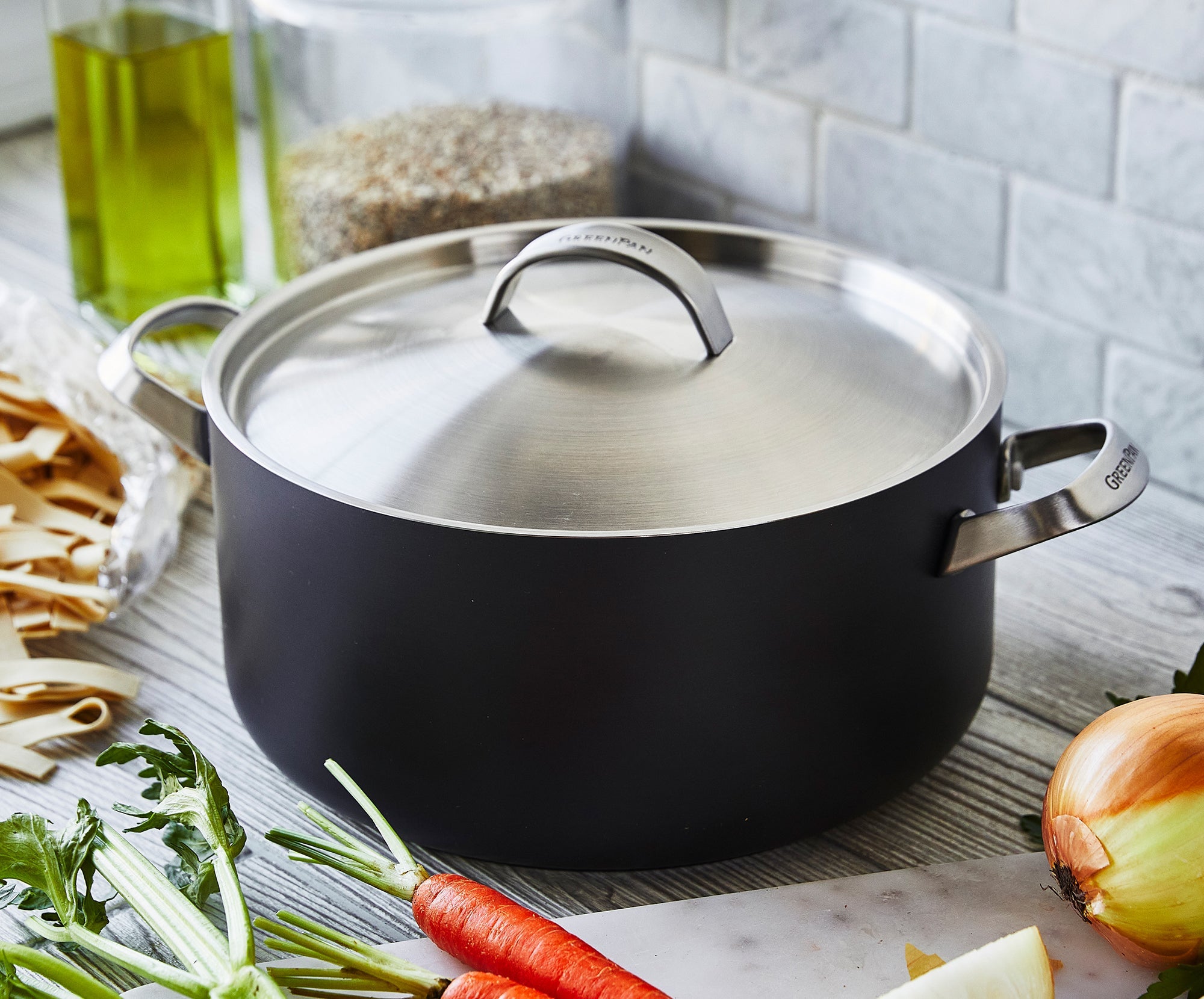 Black GreenPan pot with lid on kitchen counter, surrounded by assorted vegetables, dry pasta, and a bottle of olive oil.