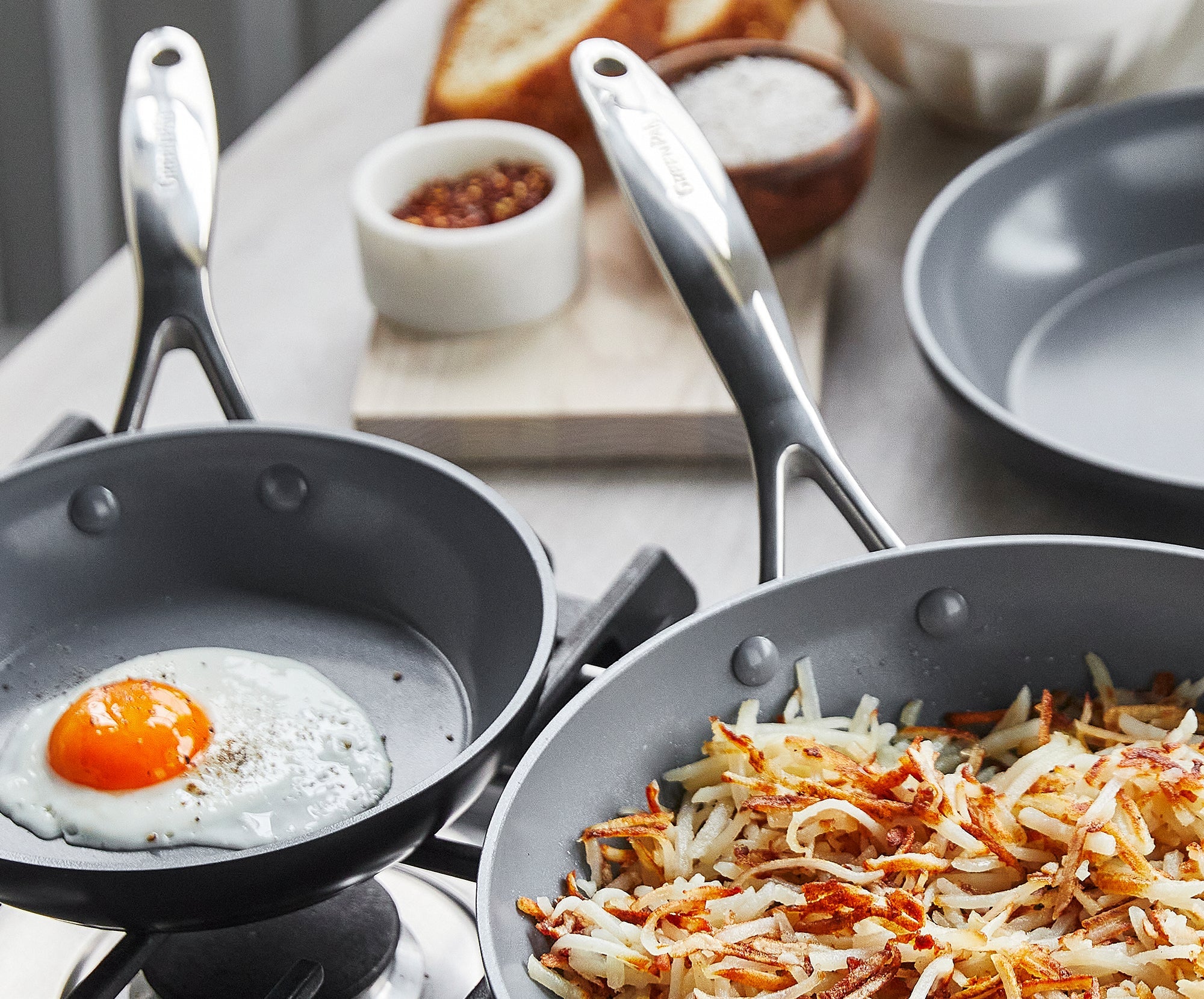 Two GreenPan ceramic nonstick skillets on a stovetop, one with a fried egg, the other with hash browns; bread and condiments behind.