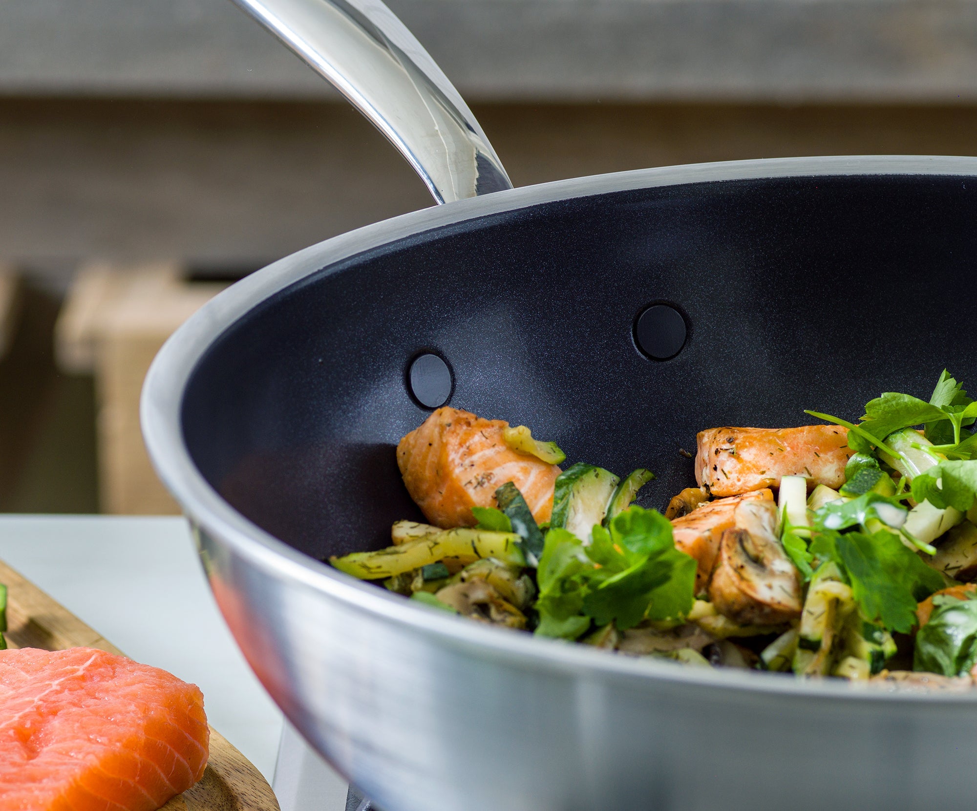 Salmon and vegetables cooking in a GreenPan ceramic pan, with raw salmon fillets on a wooden board nearby.