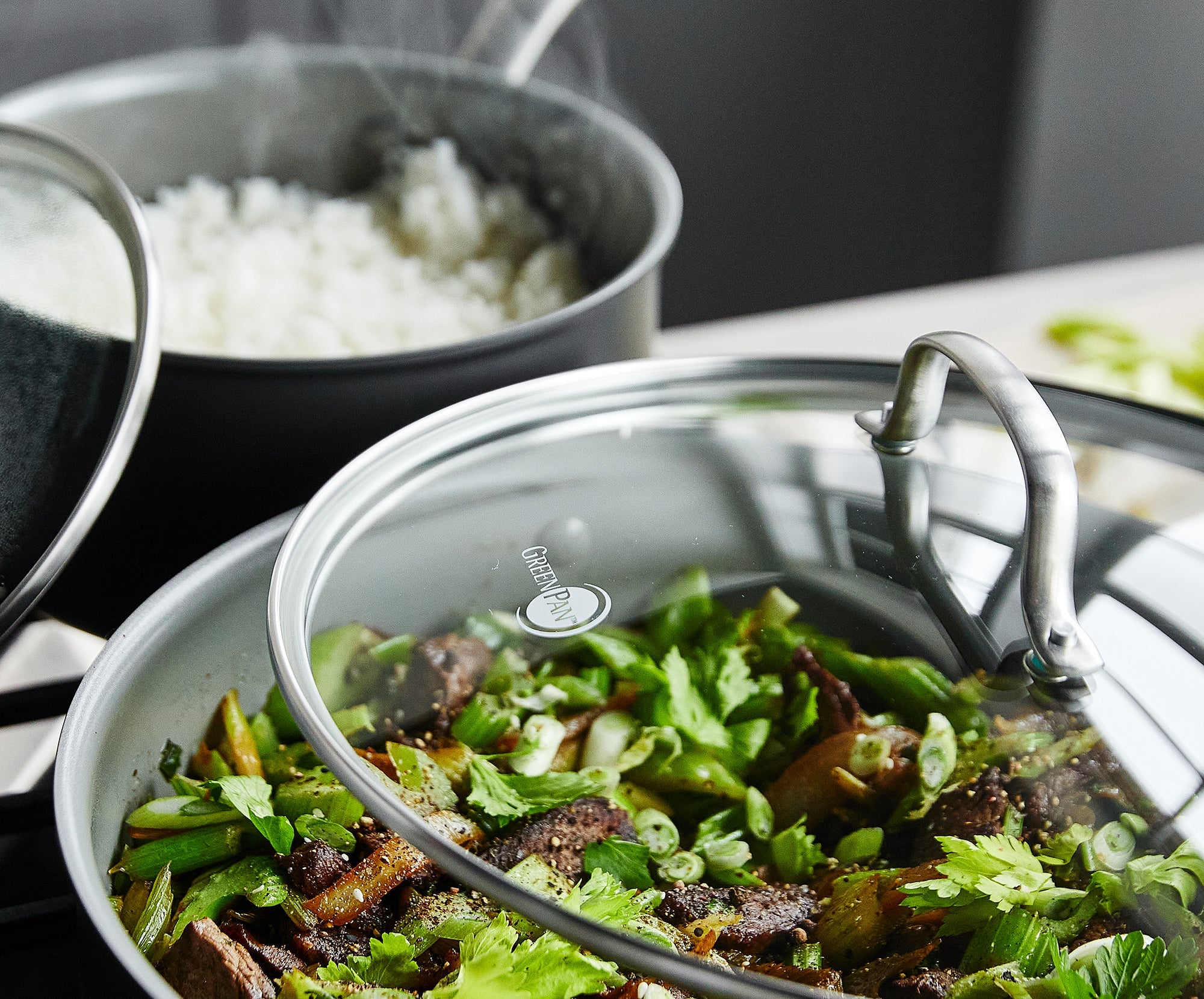 Ceramic nonstick pot with steamed white rice and covered pan with stir-fried vegetables and beef topped with herbs on a stove.