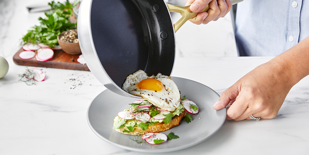 Fried egg being placed on avocado toast with radish slices and herbs, served on a gray plate.